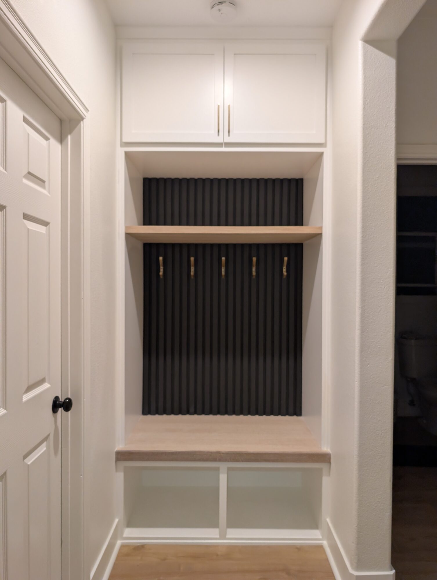 Finished mudroom with painted cubbies, black slat wall, white oak bench and five brass coat hooks, Winchester CA